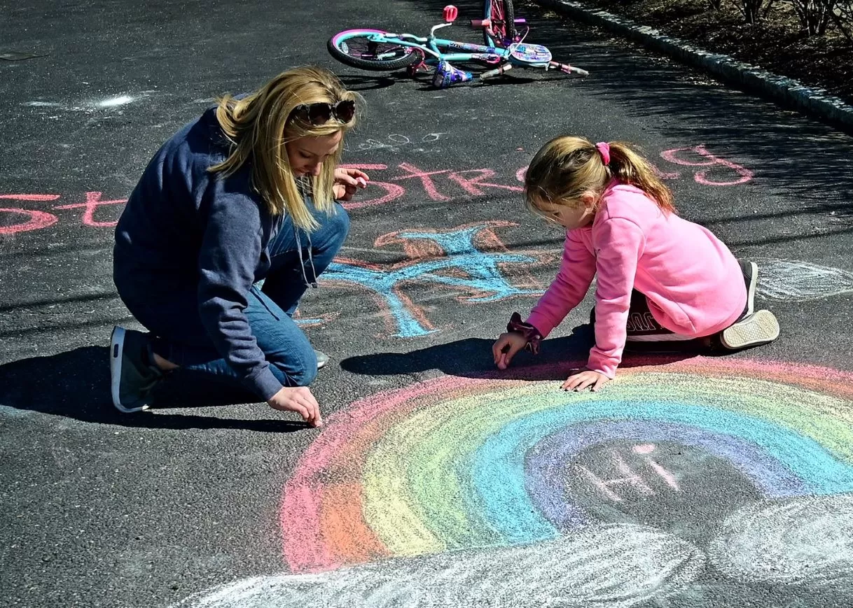 mom and daughter coloring a rainbow with sidewalk chalk on a blacktop.